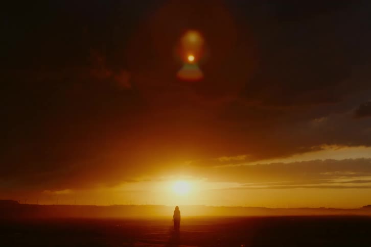 A silhouetted woman walks towards a dramatic golden sunset across a vast, empty landscape with a lens flare