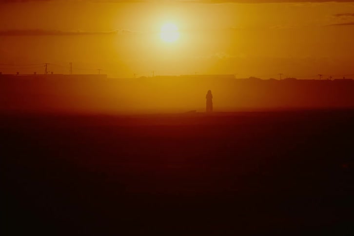 A silhouetted figure stands alone in a hazy, desolate landscape during a vibrant orange sunset
