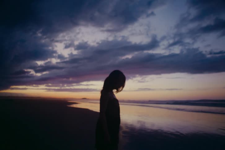 A woman's silhouette is captured as she stands on a reflective beach during a moody sunset, with the camera slowly zooming in