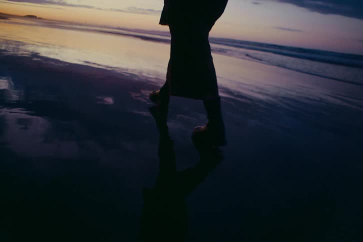 The silhouette of a person walks in slow motion along the wet sand of a beach at dusk, their reflection cast below them