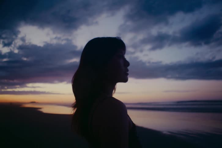A silhouetted woman stands contemplatively on a beach at dusk, looking out over the ocean