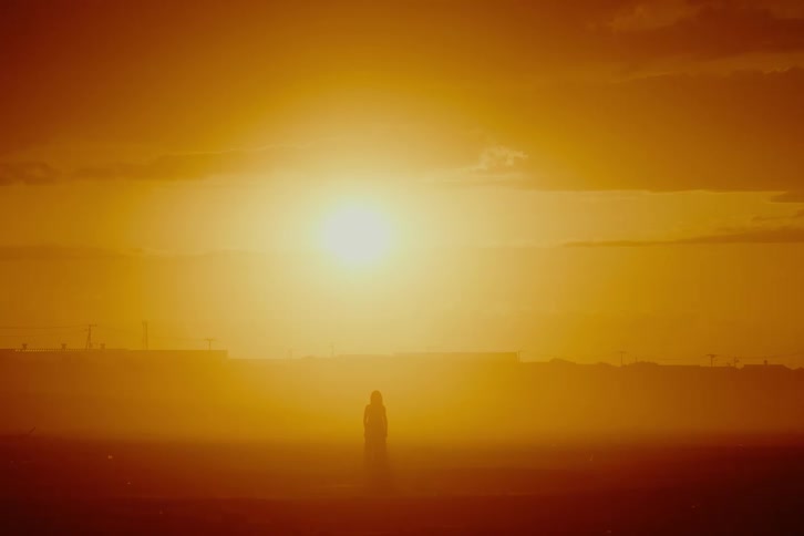 A lone person stands silhouetted against a brilliant, hazy sunset in a desolate landscape
