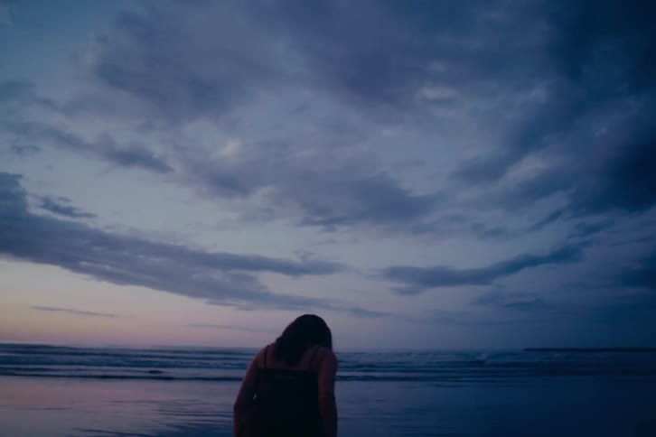 The silhouette of a young woman is seen from behind as she contemplates the ocean waves at twilight