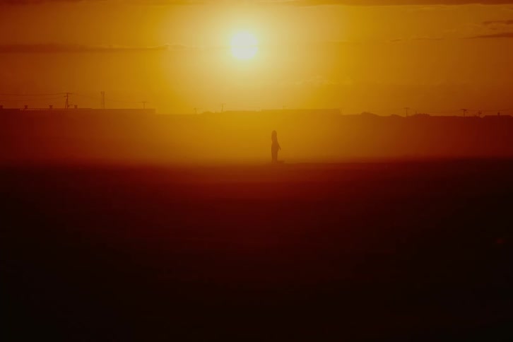 A lone figure stands silhouetted in a desolate, hazy landscape during a vibrant orange sunset