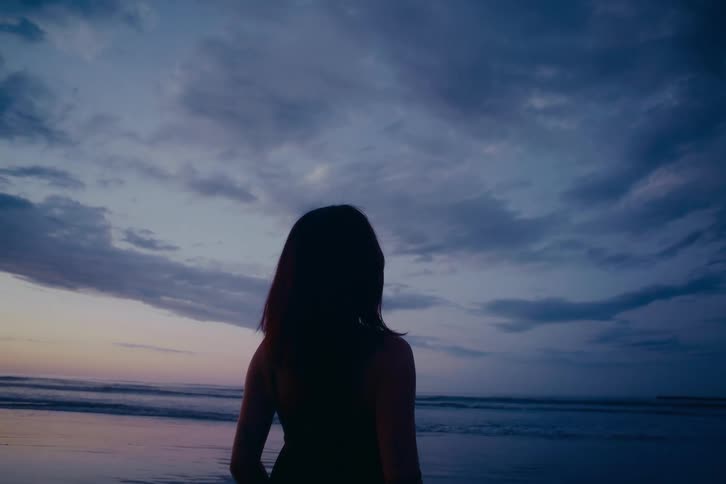 A silhouetted woman stands still on a beach, looking out at the ocean waves under a dramatic twilight sky