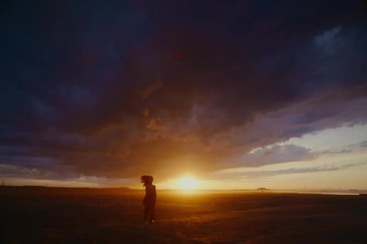 A silhouetted woman with flowing hair runs toward the camera across a vast plain under a dramatic cloudy sky at sunset