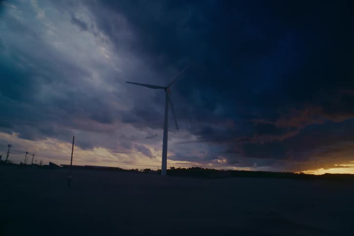 A solitary wind turbine slowly spins against a dramatic sky filled with dark, stormy clouds as the sun sets on the horizon