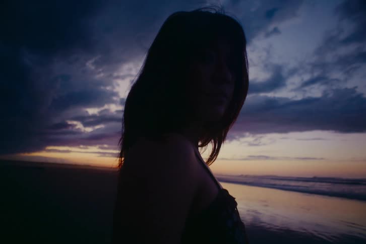 A silhouetted woman stands thoughtfully on the beach at dusk under a dramatic, cloudy sky