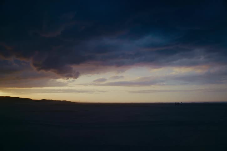 A wide, dramatic shot shows two distant figures walking on a vast beach under a moody, cloud-filled sky at twilight