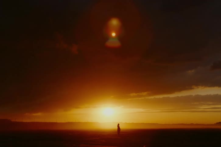 A woman in silhouette walks toward a brilliant orange sunset with dramatic dark clouds overhead