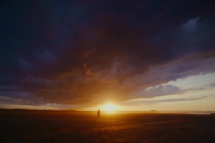 A woman runs in slow motion across a vast, empty landscape towards a dramatic golden sunset under dark, stormy clouds