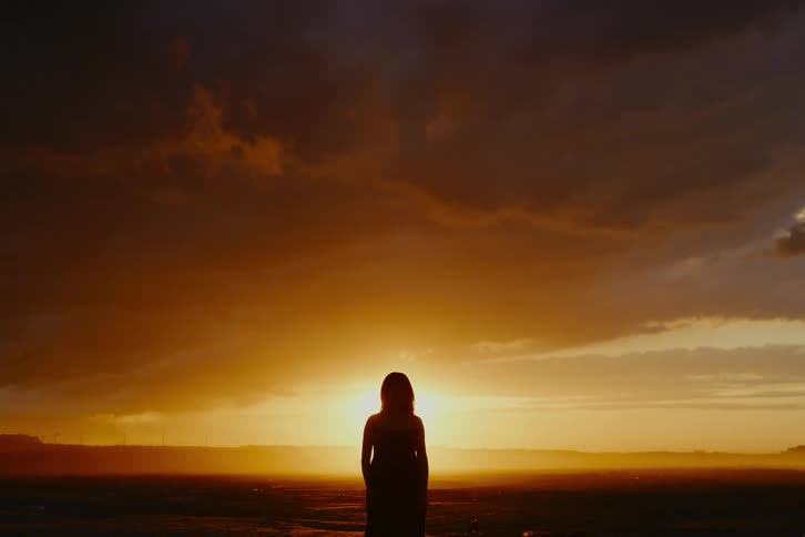 Silhouette of a woman standing alone and watching a dramatic golden sunset under dark clouds