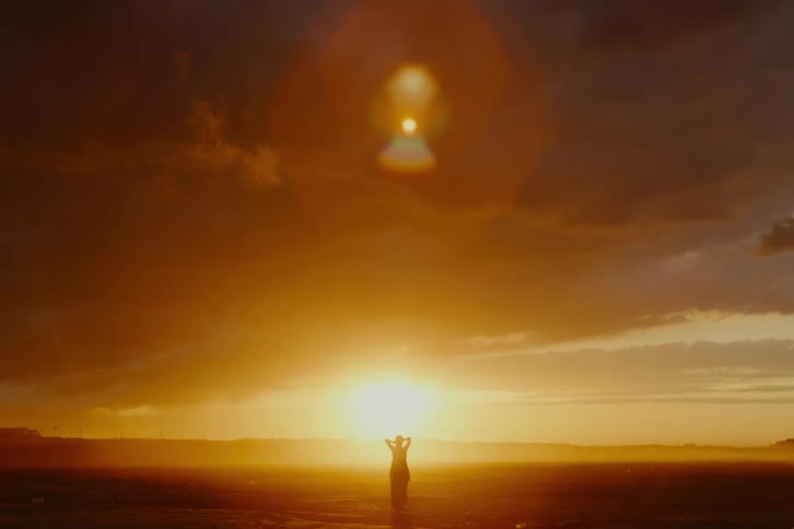 A lone woman's silhouette is framed against a vast, dramatic golden sunset in an open landscape