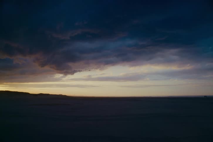 A vast, desolate beach at twilight under a dramatic, dark sky with the last light of sunset on the horizon