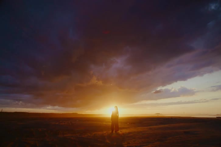 A silhouetted figure stands alone in a vast landscape, gazing at a breathtakingly dramatic and cloudy sunset on the horizon