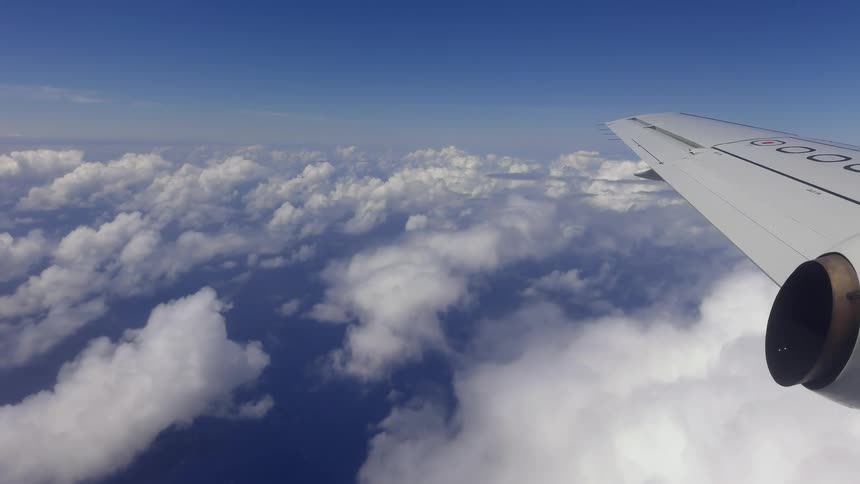 An aerial view from an airplane window shows the wing flying high above a sea of fluffy white clouds and a deep blue ocean