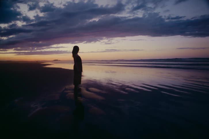 A solitary woman's silhouette is reflected in the wet sand as she stands peacefully watching the ocean waves at twilight
