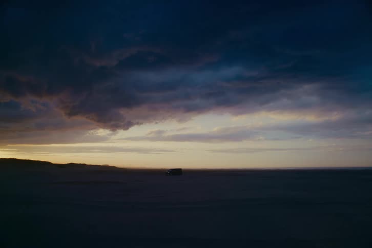 A lone van is parked on a vast, empty beach as dramatic storm clouds gather over the horizon at dusk