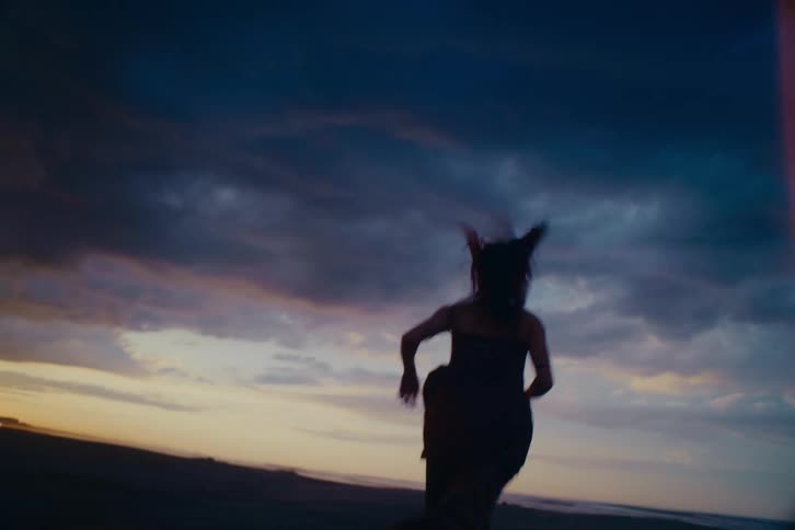 A silhouetted woman runs with purpose across a dark beach in slow motion under a dramatic and moody twilight sky