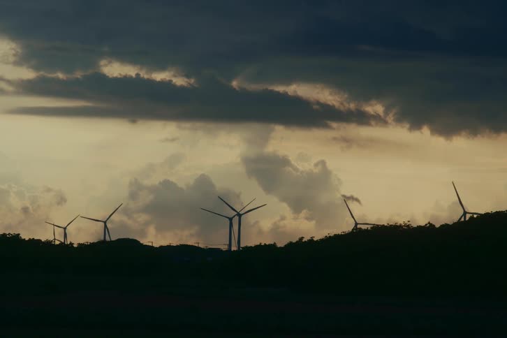 Silhouetted wind turbines on a hill spin gracefully against a moody, cloud-filled sky at dusk