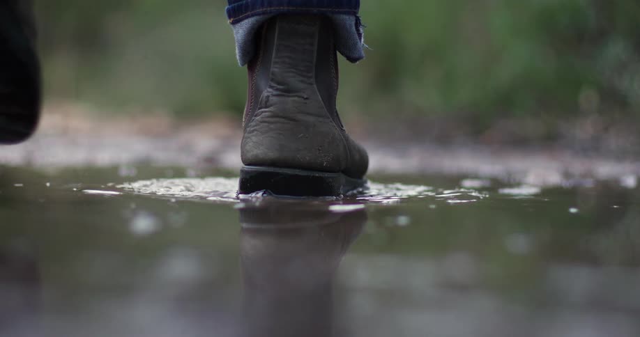 Super slow motion close-up of a person's brown boot splashing down into a muddy puddle on a trail