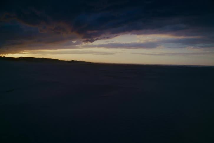 A vast, dark desert landscape sits under a moody, dramatic sky as the last light of sunset fades on the horizon