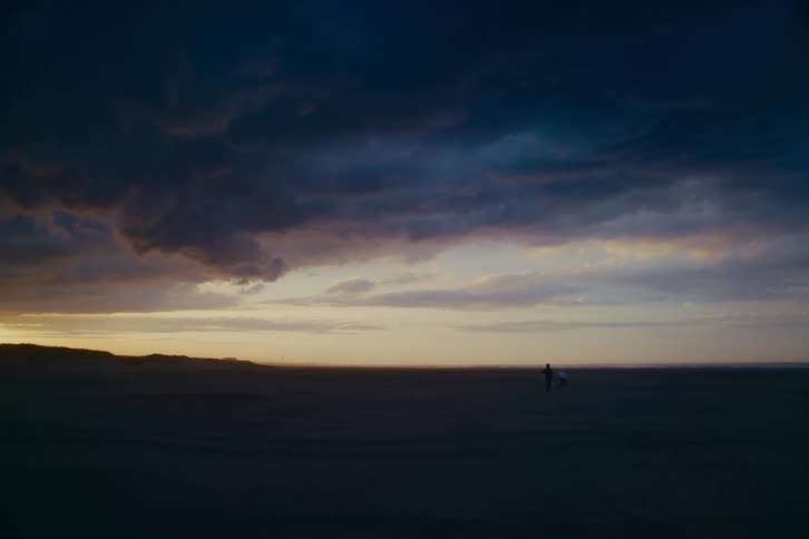 A silhouetted couple walks across a vast, empty beach under a moody, cloud-filled sky at dusk