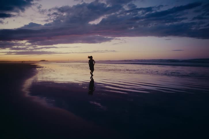 A silhouetted woman runs in slow motion along the shoreline during a dramatic purple sunset, her reflection cast on the wet sand