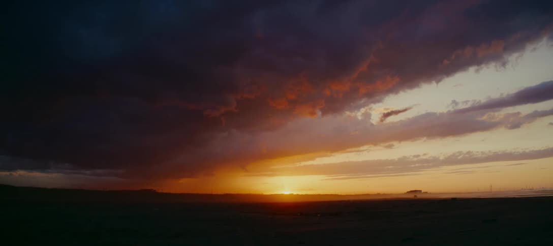 A dramatic sunrise illuminates the horizon over a vast, flat landscape as dark storm clouds gather overhead