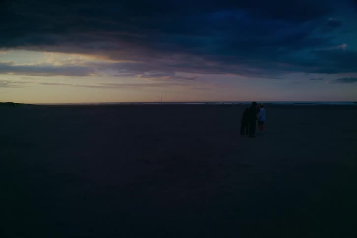 Silhouetted figures stand on a vast, empty beach under a dramatic, cloud-filled twilight sky