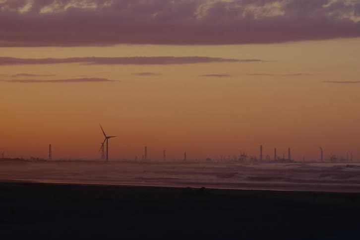 Spinning wind turbines and an industrial factory skyline are seen across a foggy ocean during a colorful sunrise