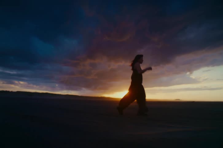 A young woman dances in slow motion in a desolate landscape with a windmill in the background during a beautiful orange sunset