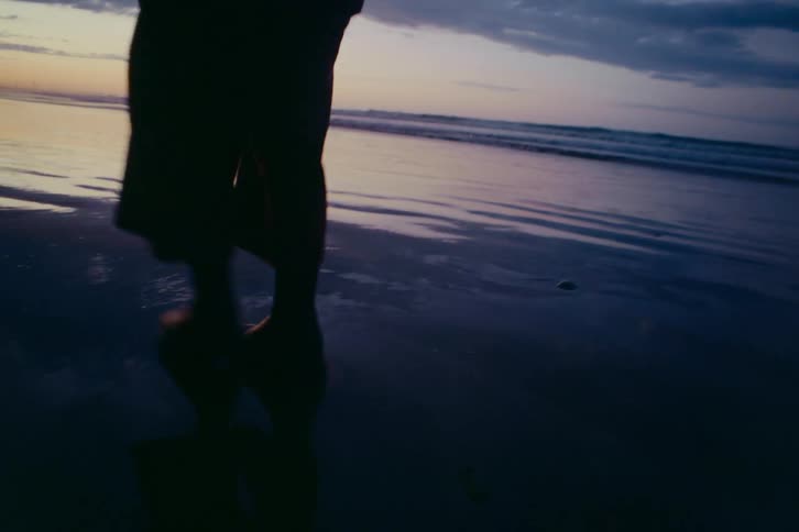 A person's silhouetted legs and feet walk in slow motion along the wet, reflective sand of a beach during a calm, moody sunset