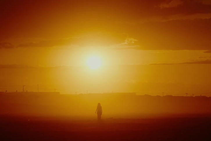 A woman stands silhouetted against a brilliant orange sunset, bathed in a hazy, atmospheric light
