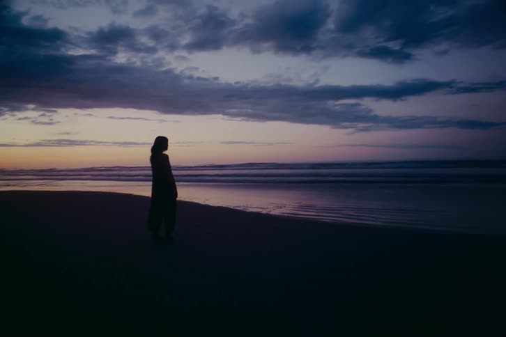 A silhouetted woman walks along the shoreline at dusk, stopping to gaze at the moody, colorful sky over the ocean