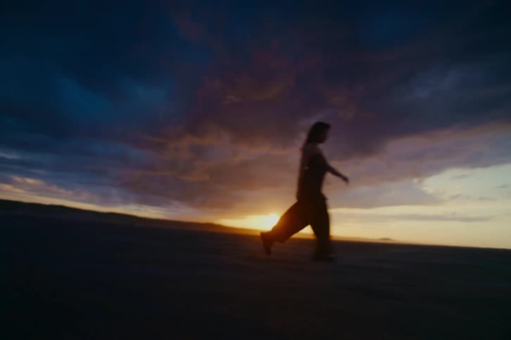 A silhouetted woman moves with expressive freedom across a barren landscape under a stunning sunset sky