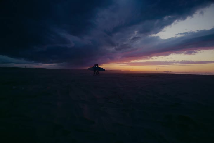A silhouette of two surfers carrying a surfboard walks across a dark beach under a dramatic sunset sky