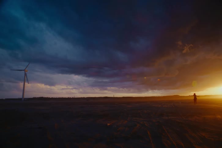 A carefree woman runs and dances through a vast, barren landscape under a stormy sunset sky with a single wind turbine on the horizon