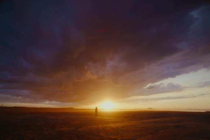 A lone figure is silhouetted against a vast desert landscape under a dramatic, cloudy sunset