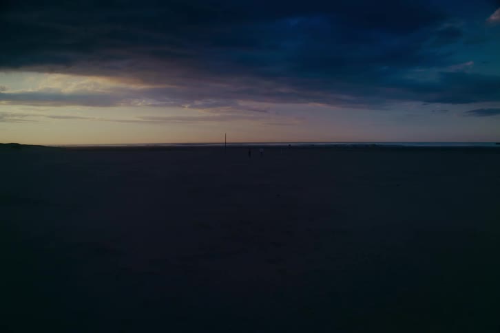 A wide, static shot of a vast, dark beach at twilight as two distant figures walk under dramatic, stormy clouds