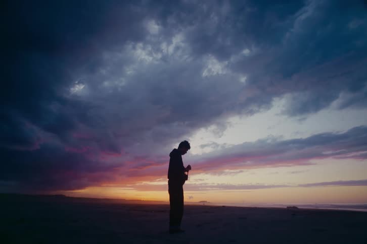 A silhouetted man stands alone on a beach at sunset, looking down at his hands under a vast, dramatic cloudy sky