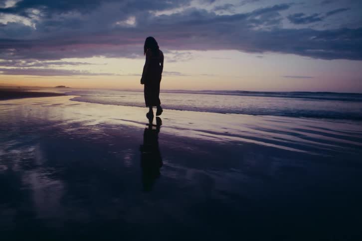 A slow-motion wide shot of a woman's silhouette walking alone on a serene beach at twilight, her reflection cast on the wet sand