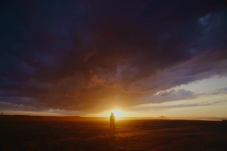 A lone figure walks in slow motion across a vast, empty landscape towards a dramatic, cloudy sunset