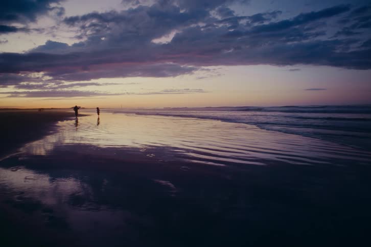 Silhouettes of a surfer and a child walking along a reflective beach during a beautiful purple sunset