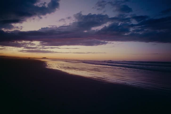 Calm waves lap the shore of a dark sand beach during a dramatic purple and blue twilight