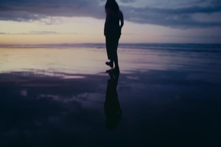 A silhouette of a woman walks in slow motion along the shoreline at sunset, her reflection cast on the wet sand
