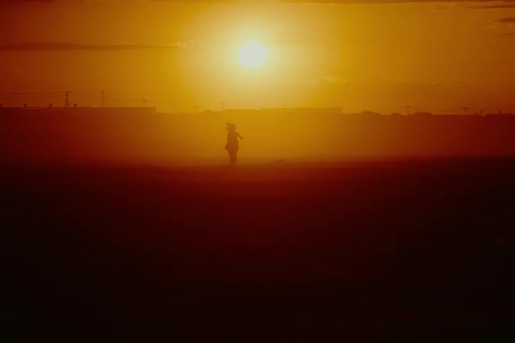 The silhouette of a woman dances slowly across a hazy field during a vibrant orange sunset