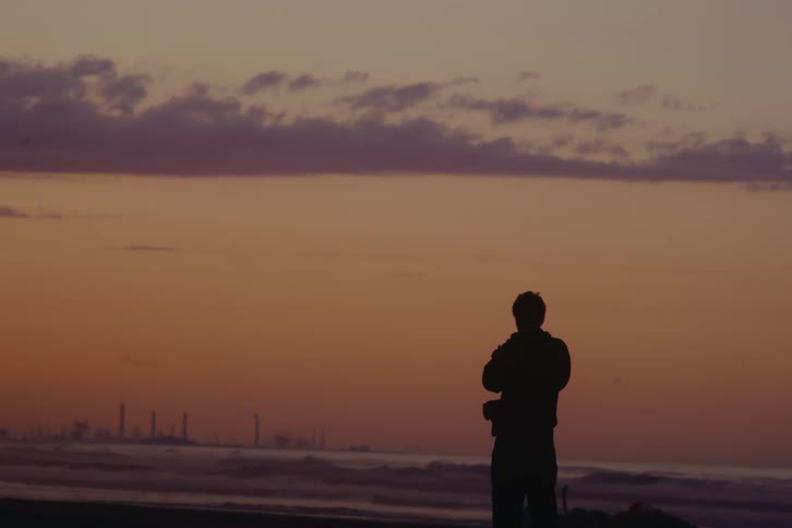 A lone photographer is silhouetted against a colorful sunrise sky while taking photos of the ocean and a distant industrial skyline