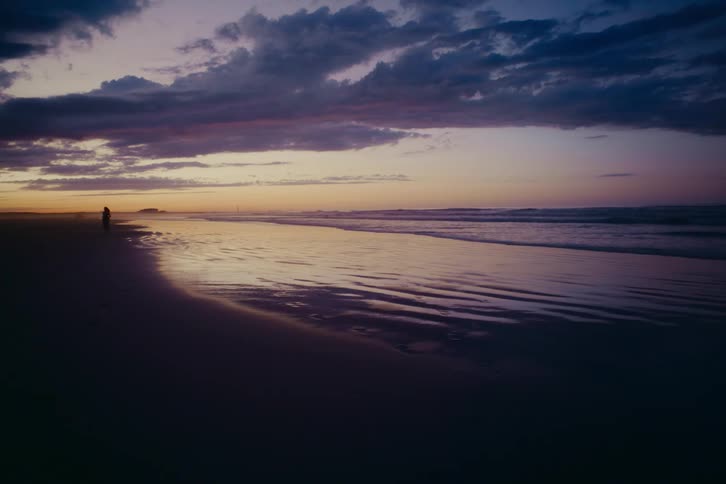 A silhouetted woman runs in slow motion towards the camera on a reflective beach during a dramatic purple and orange sunset