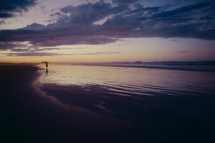 A silhouetted woman runs along a tranquil beach at dusk as the colorful sky reflects on the wet sand